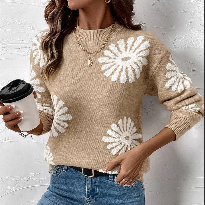Woman wearing a tan knit sweater with white daisy patterns, light blue jeans, and gold layered necklaces.