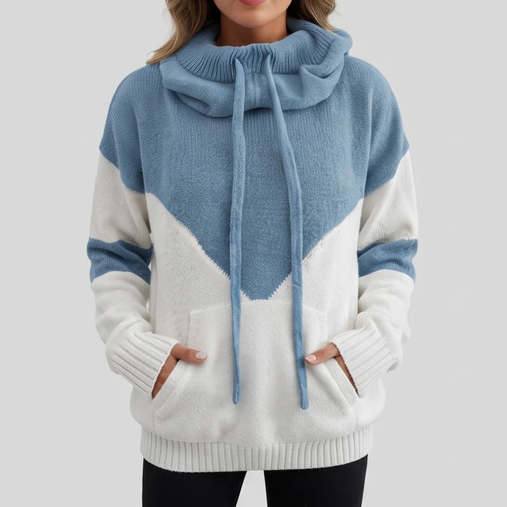 Woman wearing a blue and white color-block knit cowl neck sweater with a kangaroo pocket and her hands inside.