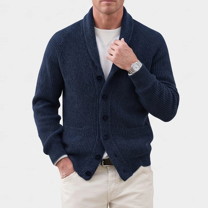 Man wearing a navy blue ribbed knit cardigan with a shawl collar, dark buttons, and front pockets, against a white background.