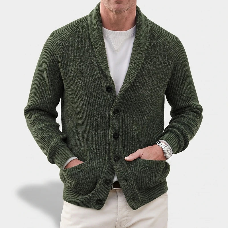 Man wearing a dark green ribbed knit shawl collar cardigan with black buttons and patch pockets, hands in pockets.