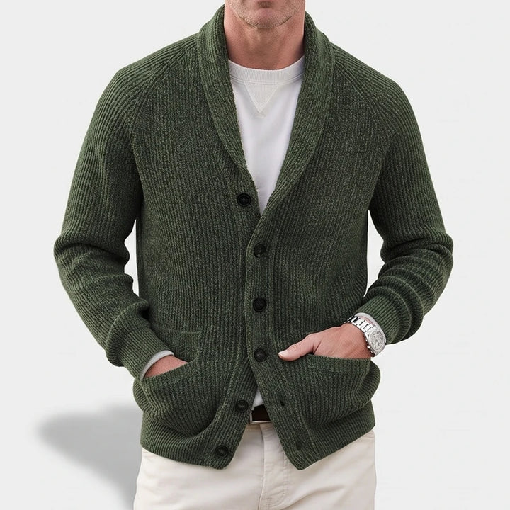 Man wearing a dark green ribbed knit shawl collar cardigan with black buttons and patch pockets, hands in pockets.