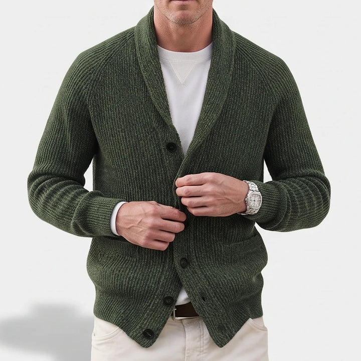 Man wearing a dark green ribbed knit cardigan with a shawl collar over a white tee, buttoning it up.
