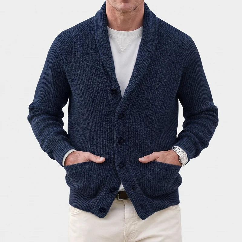 Man wearing a chunky knit navy blue cardigan with a shawl collar, front buttons, and ribbed sleeves, hands in pockets.