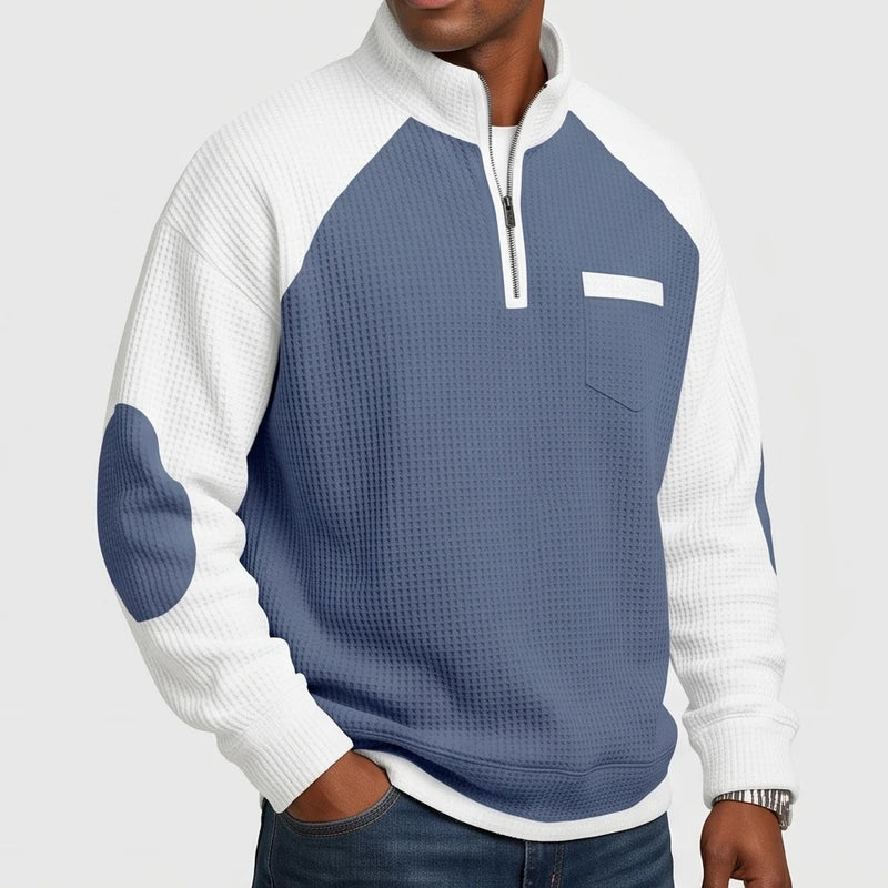 Man wearing a two-tone blue and white waffle knit sweater with a quarter-zip, chest pocket, and elbow patches.