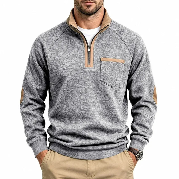 Man wearing a grey textured knit sweatshirt with a quarter-zip tan collar, tan chest pocket detail, and elbow patches.
