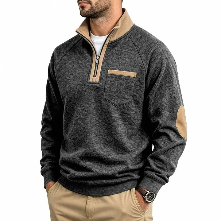 Man wearing a dark grey textured knit sweatshirt with a tan quarter-zip collar, matching elbow patches, and a chest pocket.