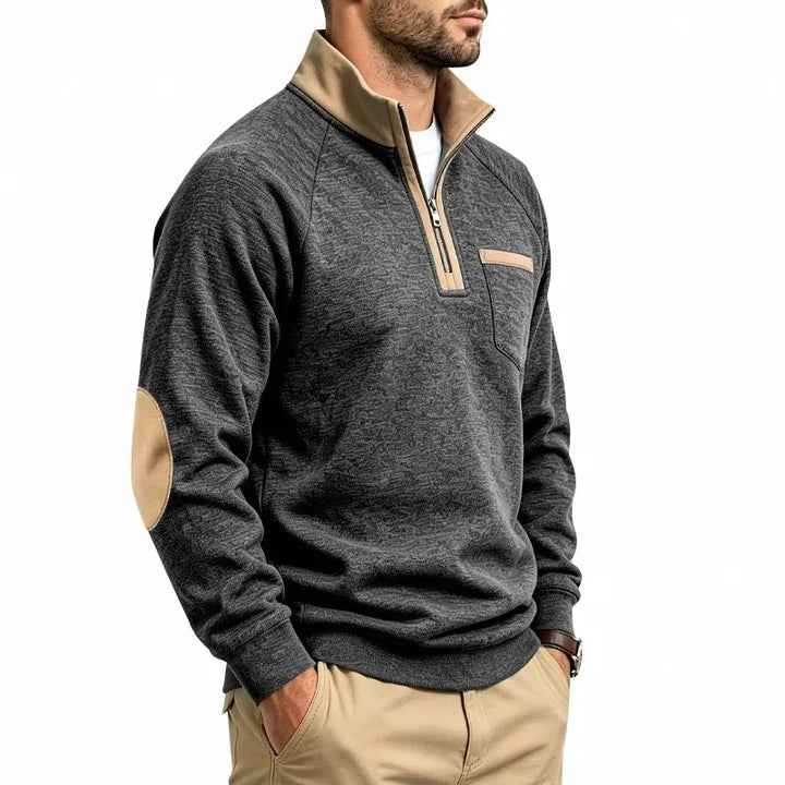 Man wearing a dark gray heathered knit sweatshirt with a beige quarter-zip collar, elbow patches, and chest pocket.
