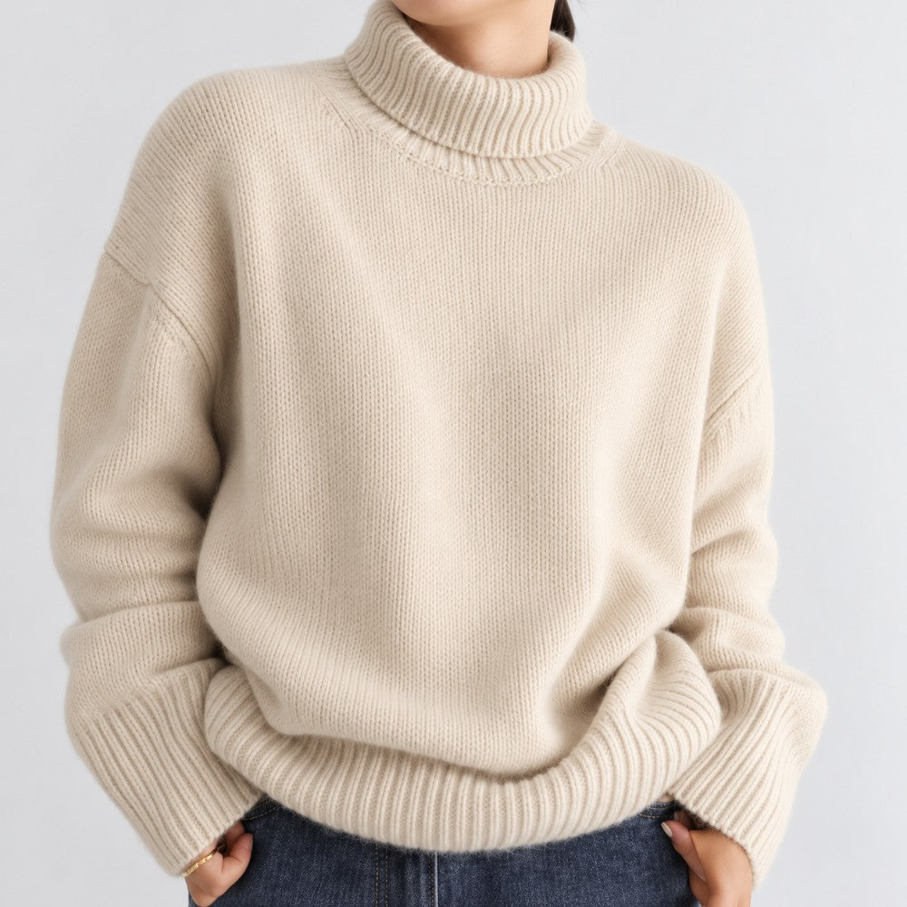 Woman wearing an oversized cream knit turtleneck sweater with ribbed trim, resting her hands in dark denim jeans.