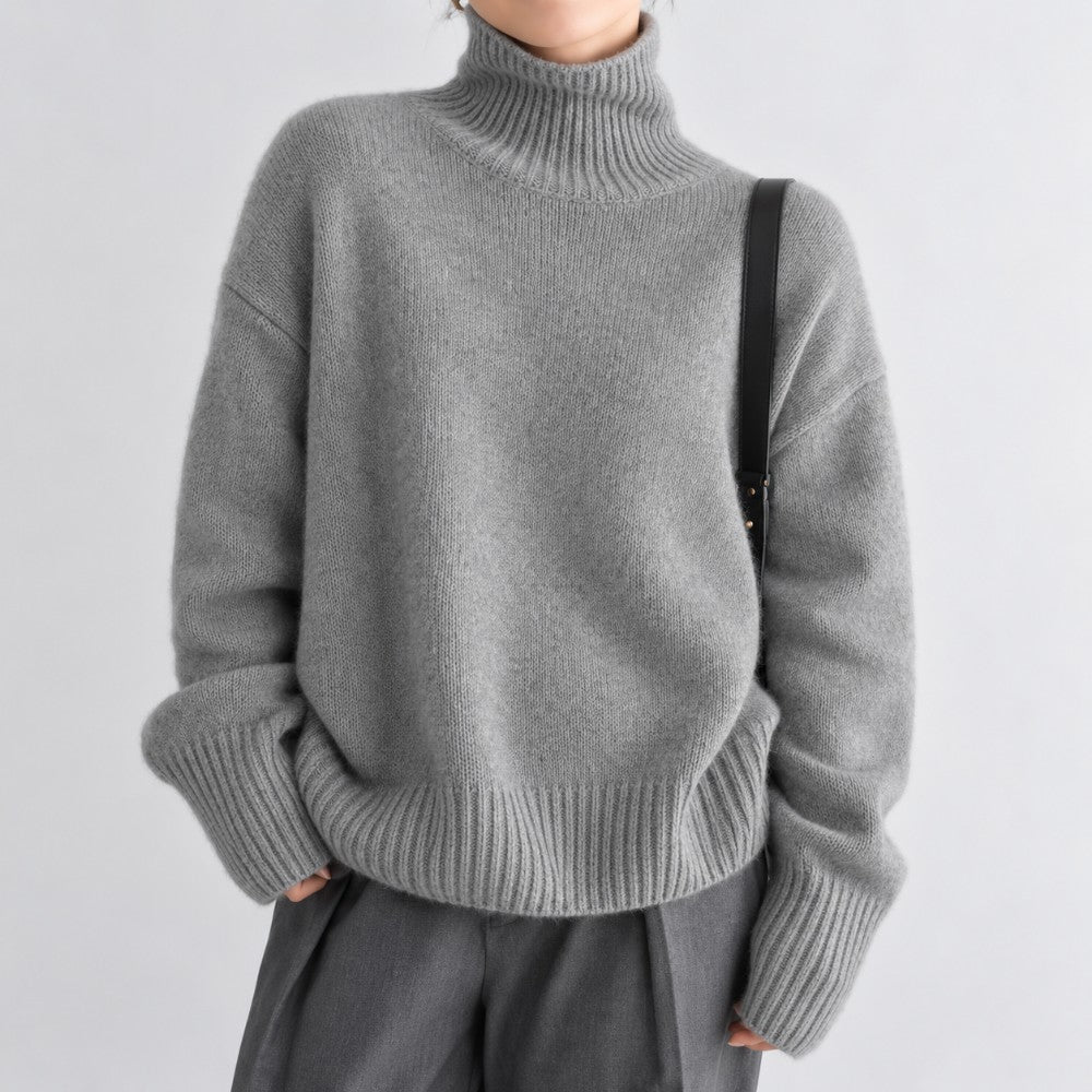 Woman wearing an oversized heather grey knit turtleneck sweater with wide ribbed cuffs and hem, against a plain white background.