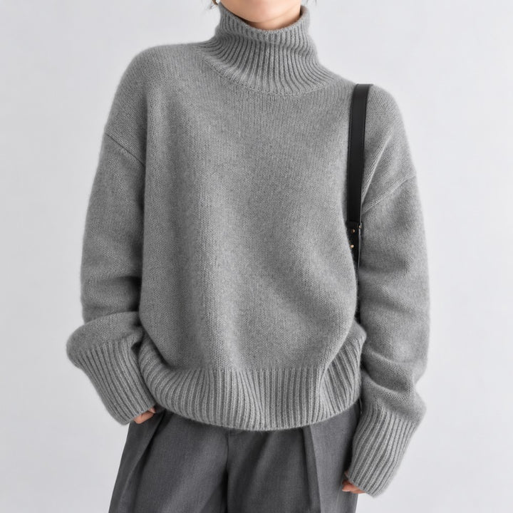 Woman wearing an oversized heather grey knit turtleneck sweater with wide ribbed cuffs and hem, against a plain white background.