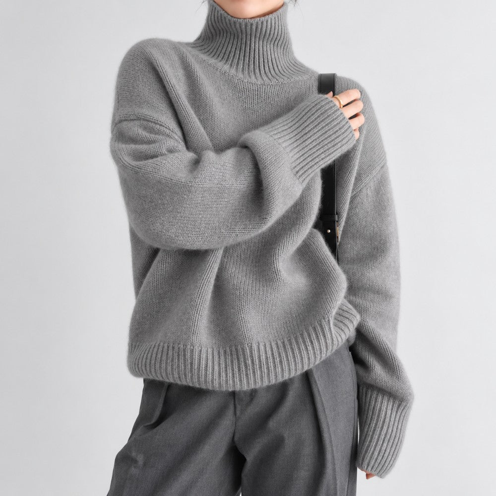Woman wearing an oversized grey knit turtleneck sweater with ribbed cuffs and matching pleated trousers against a white background.
