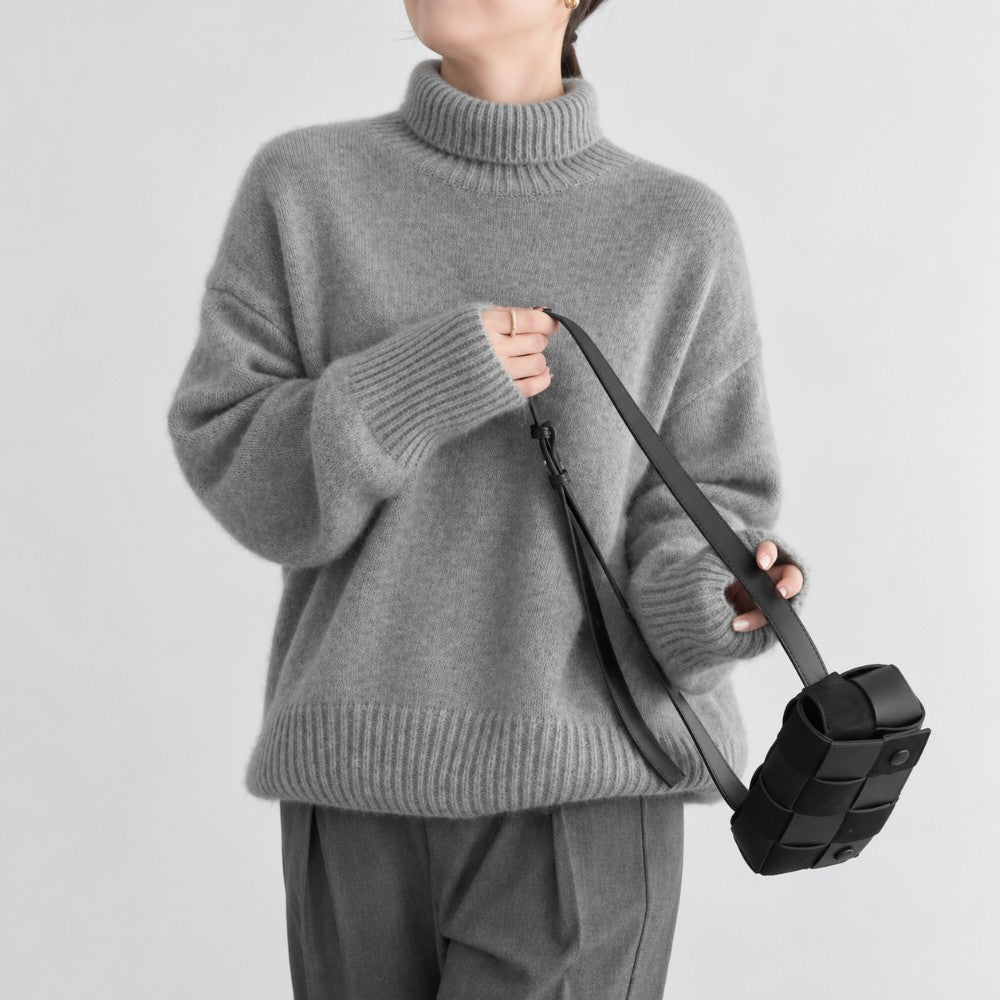 Woman wearing an oversized gray knit turtleneck sweater with ribbed cuffs and hem, holding a small black woven bag.