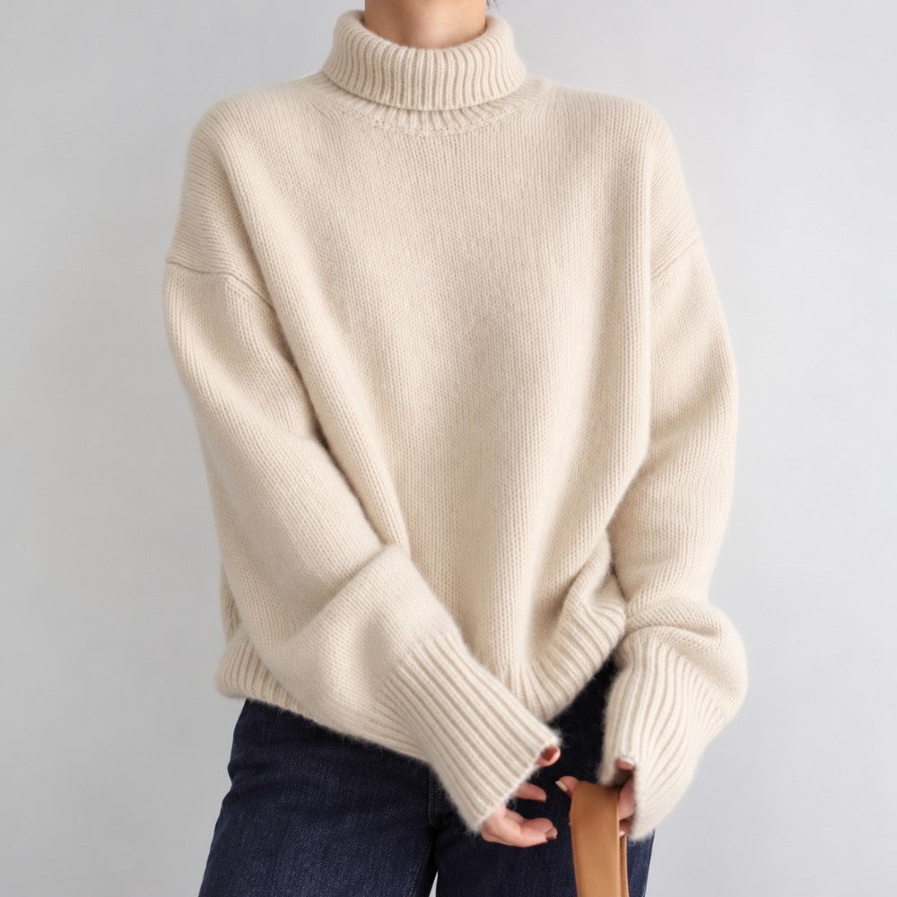 Woman wearing an oversized cream knit turtleneck sweater with dropped shoulders and wide ribbed cuffs over dark blue jeans.