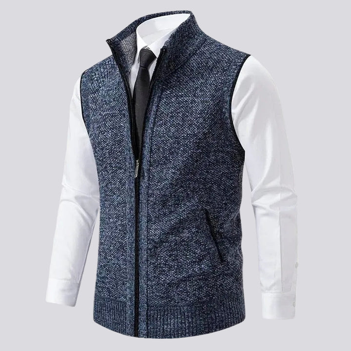 Front view of a dark blue melange knit vest with a black zipper, ribbed hem, and a left hand pocket, worn over a white shirt and black tie.