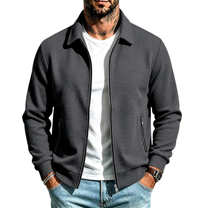 Man wearing a charcoal grey textured knit zip-up jacket with a collar and zippered pockets, hands in light wash jeans.