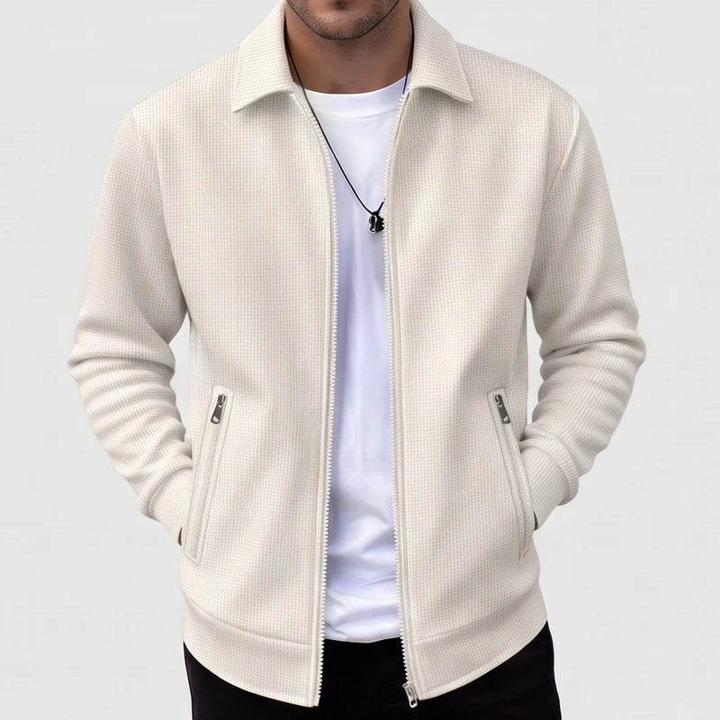 Man wearing a cream-colored waffle-knit zip-up jacket with side pockets and a white undershirt, hands in pockets.