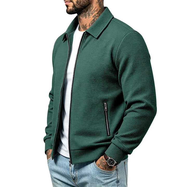 Man wearing a dark green waffle-knit zip-up jacket with a collar, black zippers, and light wash jeans.