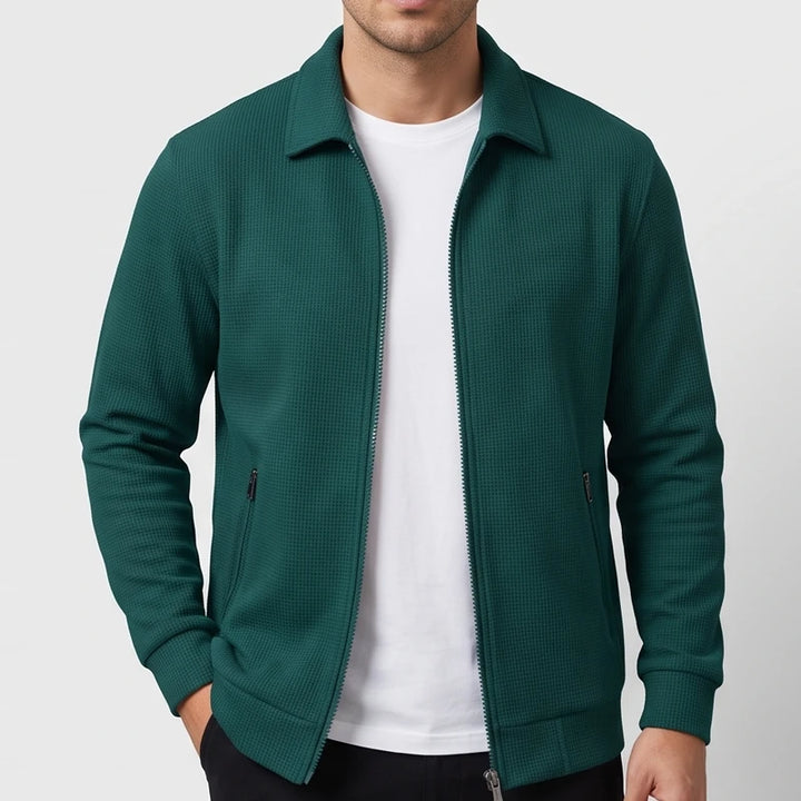 Man wearing a dark green waffle knit jacket with a collar, silver full zip, and two zippered front pockets.