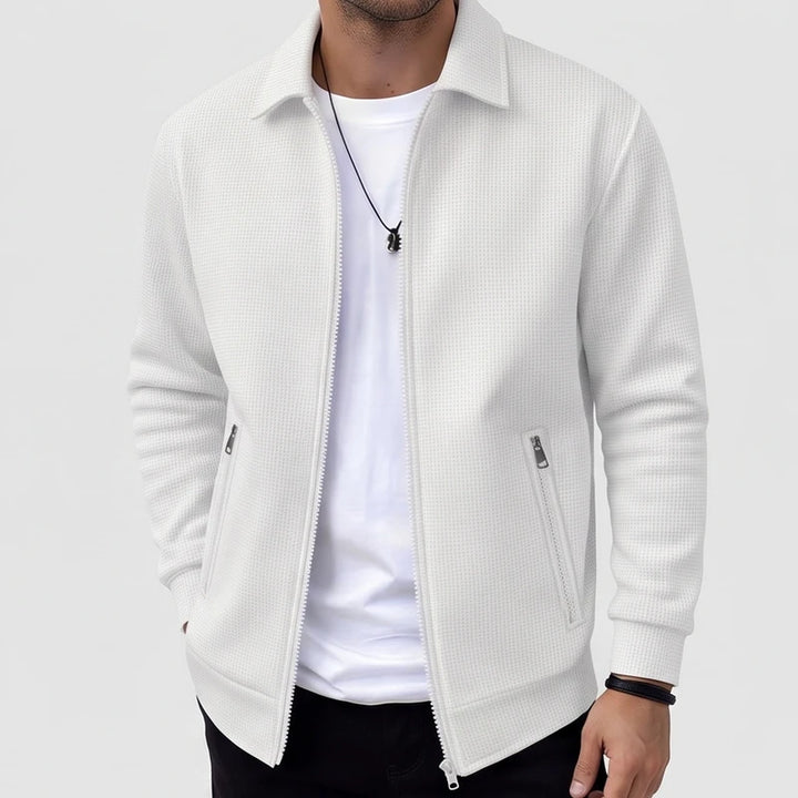 Man wearing an open white waffle-knit jacket with a collar, showing a white t-shirt and black necklace underneath.