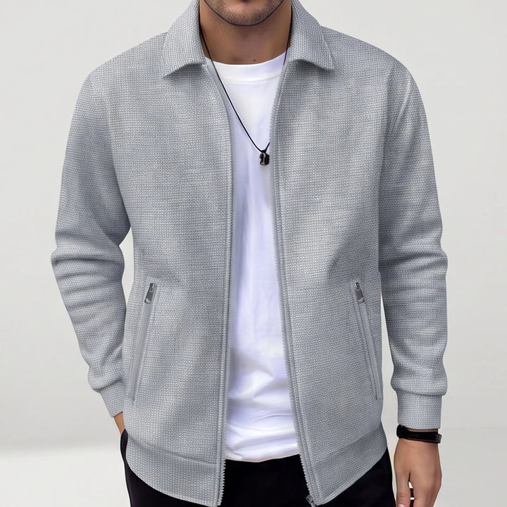 Man wearing an open light grey knit zip-up jacket with a collar, two zippered pockets, and a white t-shirt underneath.