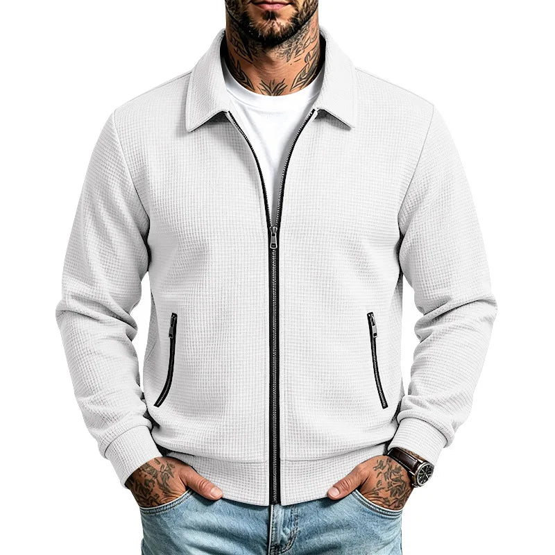 Man wearing a white waffle knit zip-up jacket with black zippers, a collar, and zippered pockets, hands in light blue jeans.