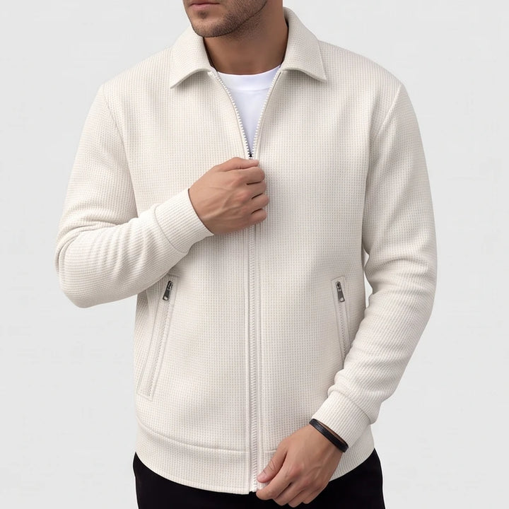 Man wearing a cream waffle-knit jacket with a collar, full zipper, and two zippered pockets, adjusting the zipper.