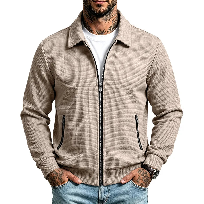 Man wearing a beige, textured zip-up jacket with a white t-shirt, light jeans, and hands in pockets.