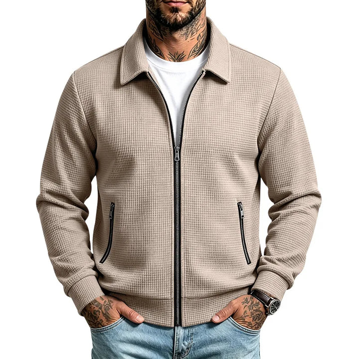 Man wearing a beige, textured zip-up jacket with a white t-shirt, light jeans, and hands in pockets.