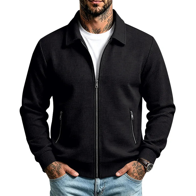 Man wearing a black textured knit zip-up jacket over a white tee, with hands in jeans pockets.