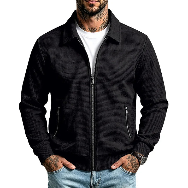 Man wearing a black textured knit zip-up jacket over a white tee, with hands in jeans pockets.