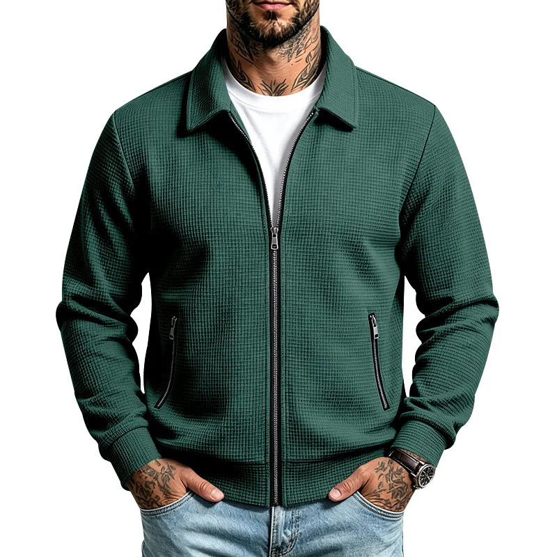 Man wearing a deep teal knit zip-up jacket with waffle texture, silver zippers, and a classic collar, hands in jeans pockets.
