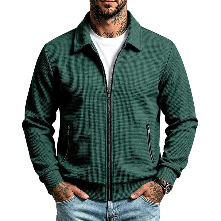 Man wearing a deep teal knit zip-up jacket with waffle texture, silver zippers, and a classic collar, hands in jeans pockets.