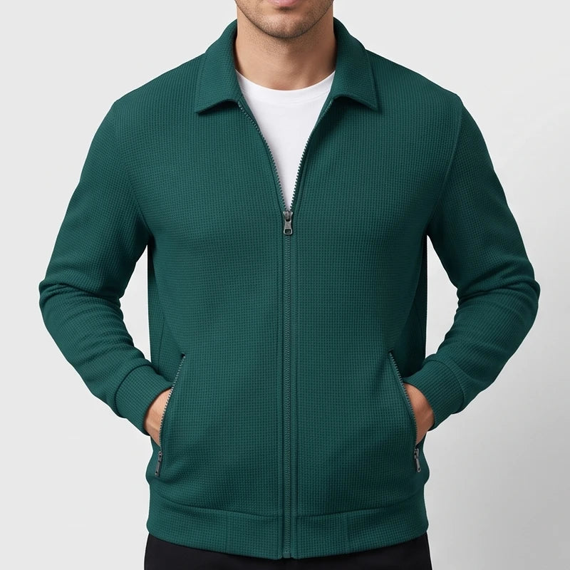 Man wearing a deep teal waffle-knit zip-up jacket with a collar, hands in front pockets, over a white shirt.