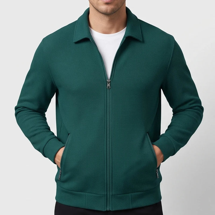 Man wearing a deep teal waffle-knit zip-up jacket with a collar, hands in front pockets, over a white shirt.