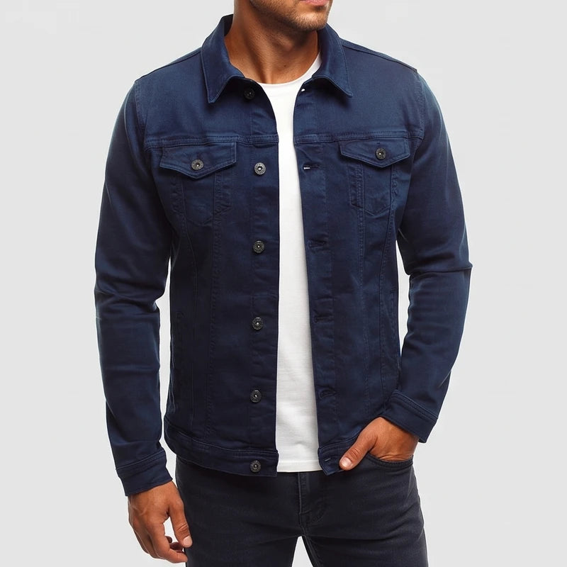 Man wearing a dark blue denim jacket with front buttons and chest pockets, over a white t-shirt and dark pants.