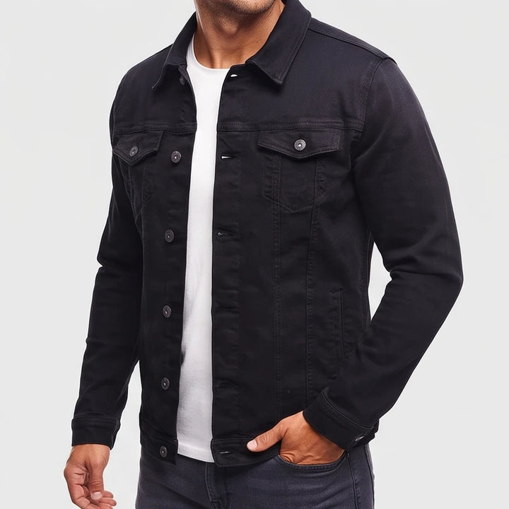 Man wearing a black denim jacket with two chest pockets and metal buttons, his hand in a dark pant pocket.