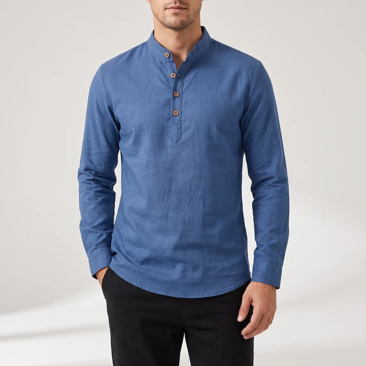 Man wearing a blue long-sleeve linen shirt with a band collar and wooden buttons, one hand in a black pants pocket.