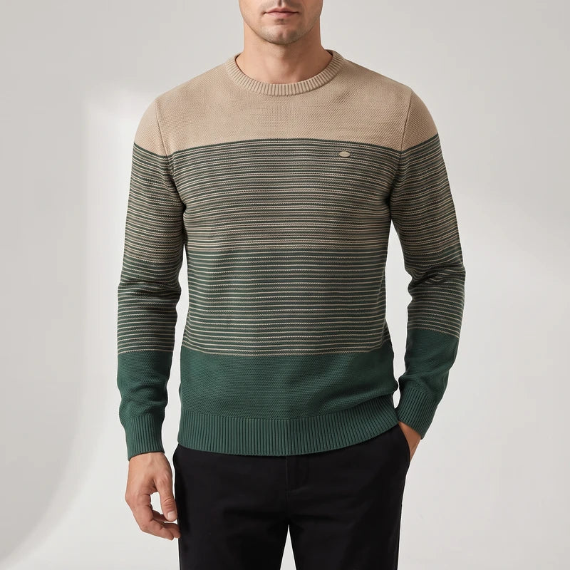 Man wearing a crewneck sweater with a solid beige top and horizontal green and beige stripes, paired with black pants.