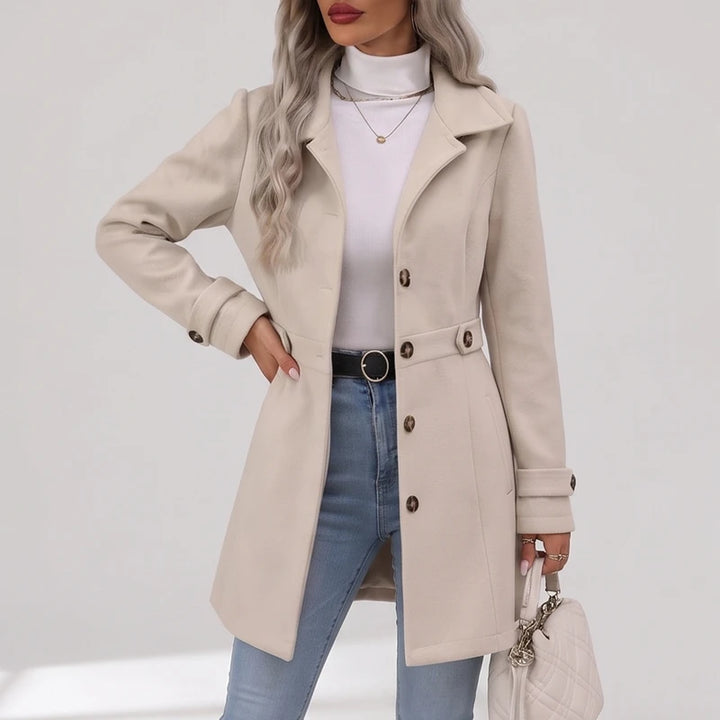 Woman wearing a light beige coat with a classic collar, large brown buttons, and belted waist details, paired with light blue jeans.