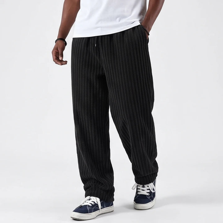 Man wearing black baggy corduroy sweatpants with a visible drawstring waist, paired with blue and white sneakers.