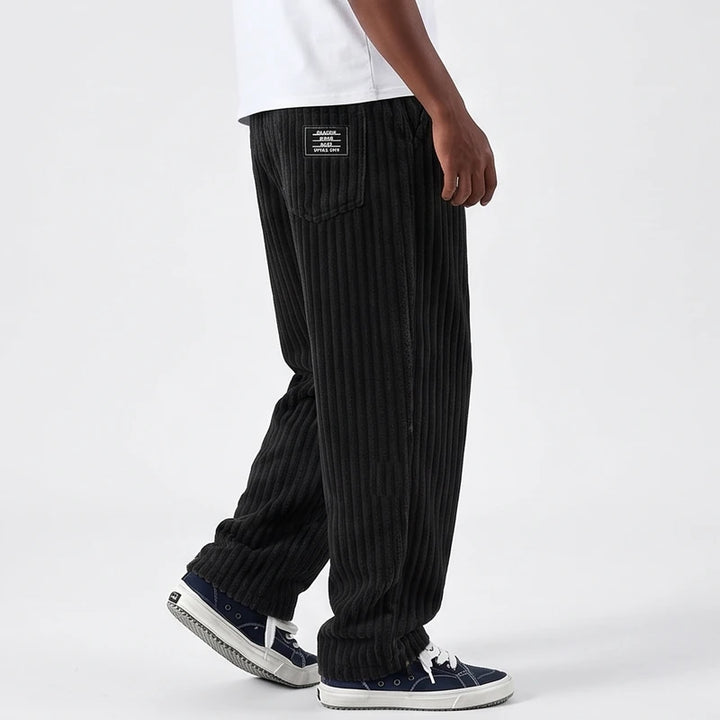 Man wearing baggy black corduroy pants with a stitched label on the back pocket, walking on a white background.