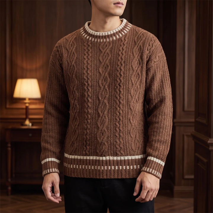 Man wearing a brown cable-knit sweater with white striped ribbed collar, cuffs, and hem, in a dimly lit room.