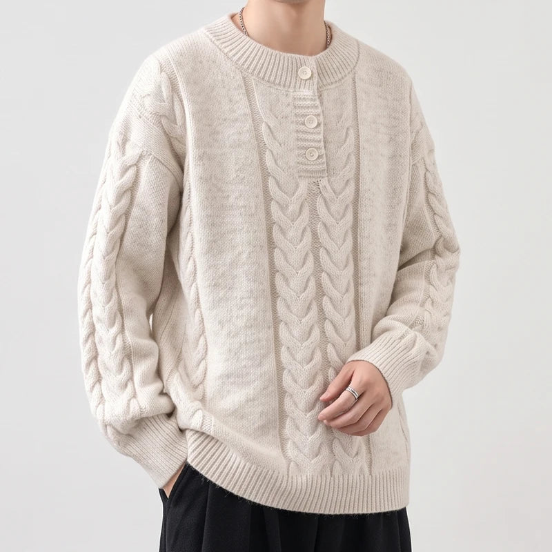 Man wearing an oversized cream cable-knit sweater with a three-button placket, hands in black pants pockets, against a white background.