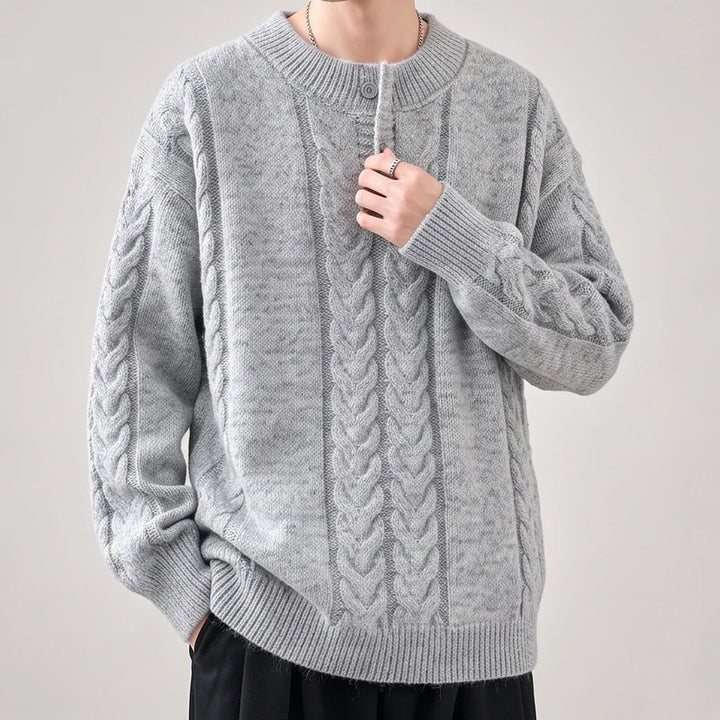 Man wearing a light gray cable-knit sweater with a ribbed crew neck and a single button placket, holding the fabric.