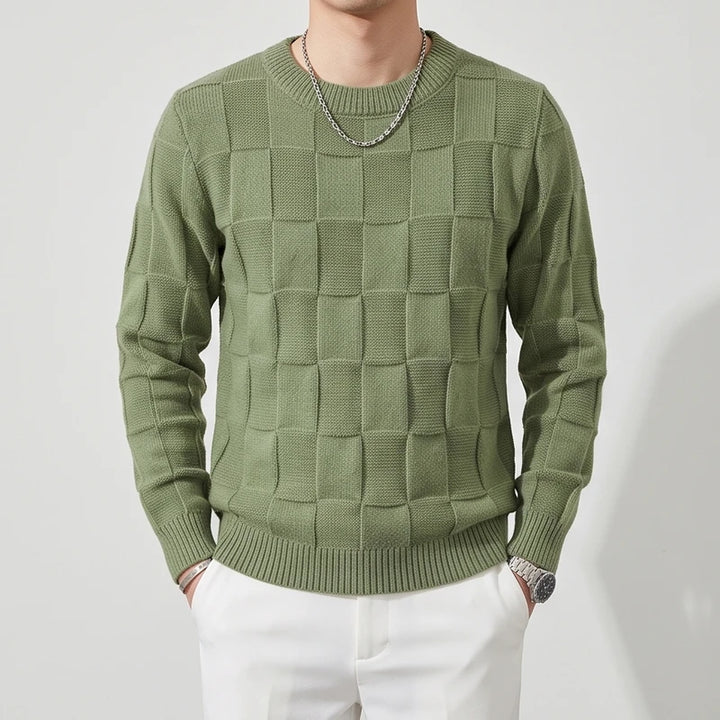 Man wearing a textured olive green knit crew neck sweater with a checkerboard pattern and white pants.