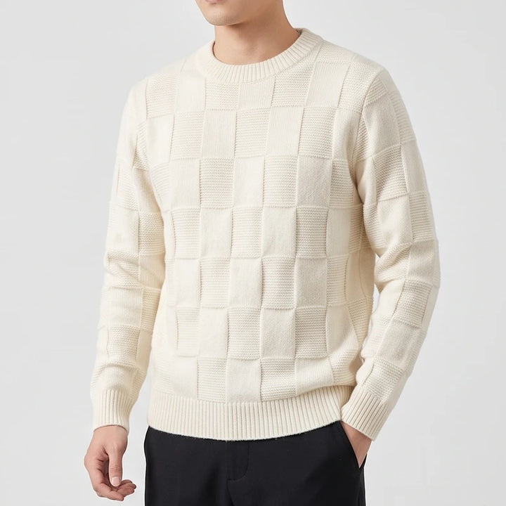 Man wearing a cream-colored, textured knit crew neck sweater with a checkerboard pattern, one hand in his black pants pocket.