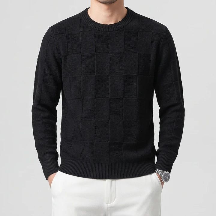 Man wearing a black checkered knit crew neck sweater and white pants, hands in pockets.