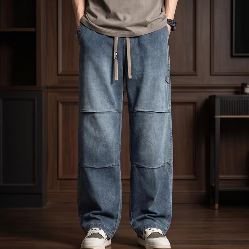 Man wearing wide-leg faded blue denim jeans with a drawstring waist, patch details, and white sneakers, standing indoors.