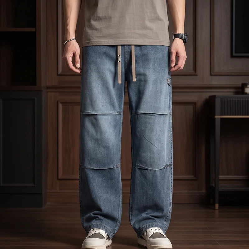 Man wearing blue wide-leg jeans with a patchwork design, drawstring waist, and carpenter loop, standing on a wooden floor.
