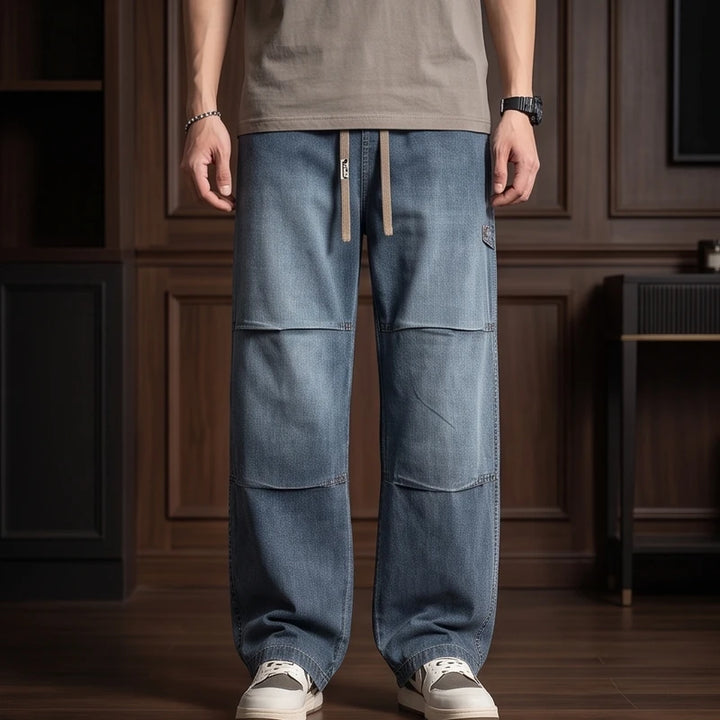 Man wearing blue wide-leg jeans with a patchwork design, drawstring waist, and carpenter loop, standing on a wooden floor.